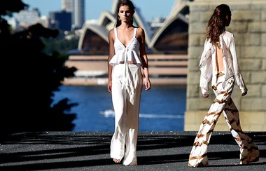 Two models walking in white outfits at an outdoor fashion show, with Sydney Opera House in the background.