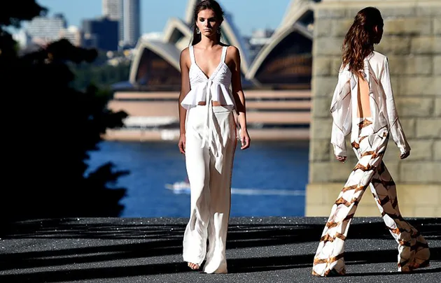 Two models walking in white outfits at an outdoor fashion show, with Sydney Opera House in the background.