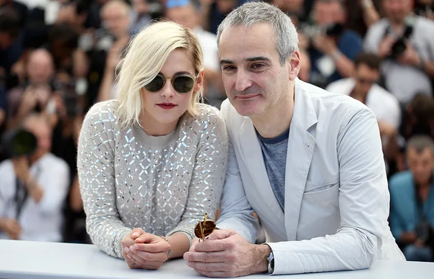 Two people at an event, one in sunglasses with light hair, the other with gray hair, surrounded by photographers.