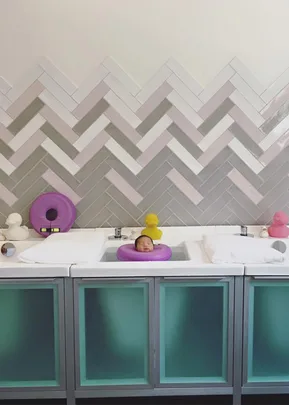 Baby in purple neck float in a baby spa bath surrounded by ducks, with herringbone pattern wall tiles.