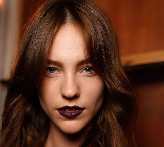 A person with long brown hair and dark lipstick looks directly at the camera, in a warm-toned indoor setting.