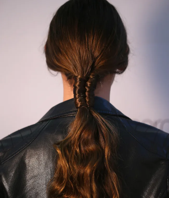 Back view of a person with long, wavy brown hair styled in a twisted ponytail, wearing a black leather jacket.