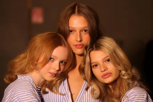 Three women with varied hair colors, wearing striped shirts, pose closely together against a dark background.