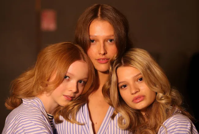 Three women with varied hair colors, wearing striped shirts, pose closely together against a dark background.