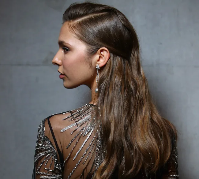 A woman with long brown hair in an elegant shiny dress and dangling earrings, looking to the left against a neutral background.