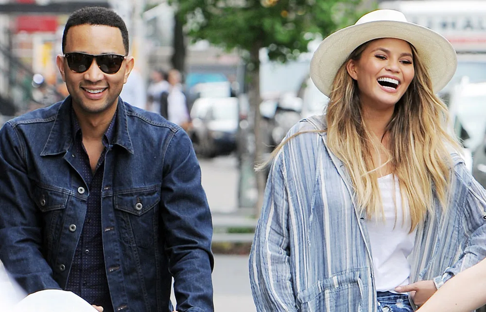 Two people smiling outdoors; one wearing sunglasses and a denim jacket, the other in a striped shirt and wide-brimmed hat.
