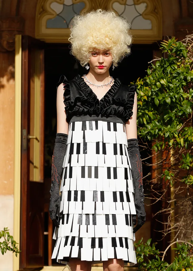 Person in a piano-themed dress with black gloves and a voluminous curly wig, standing outdoors in front of a doorway.