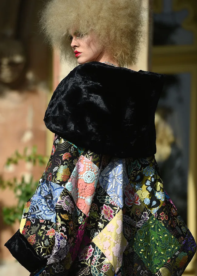 Model in a colorful embroidered patchwork coat with voluminous curly hair, posing outdoors against a blurred background.