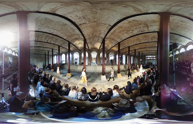 360-degree view of a fashion show with an audience seated around a runway in an industrial-style venue.
