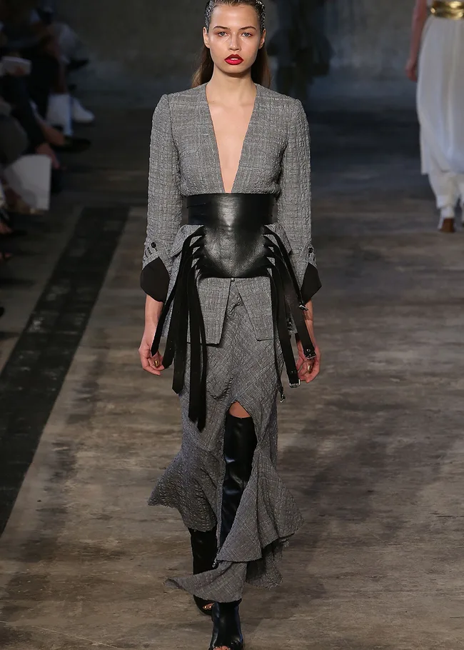Model in a gray dress with a deep neckline, leather belt with fringes, and knee-high black boots on a runway.