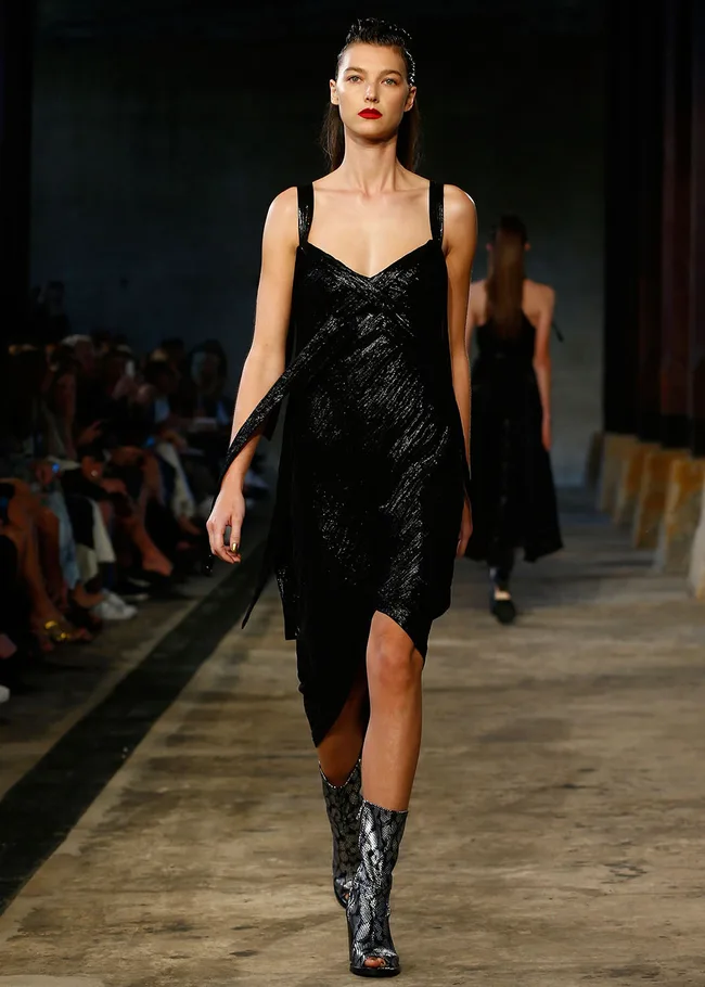 Model walking down the runway in a black dress and snakeskin boots at a fashion show.