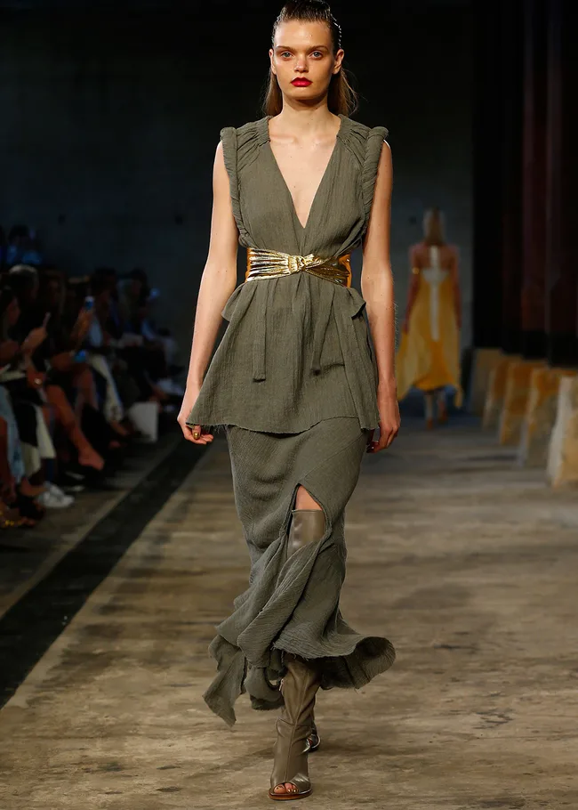 Model in a textured green dress with a gold belt and taupe boots walks the runway at a fashion show.