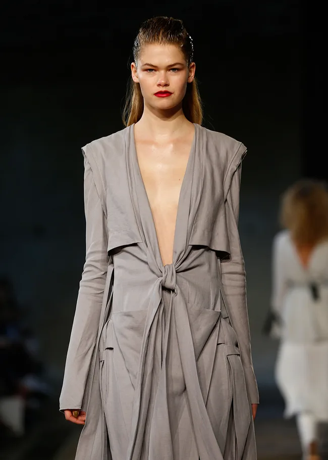Model walks runway in gray deep V-neck dress with knot detail, slicked-back hair, and red lipstick. Fashion show setting.