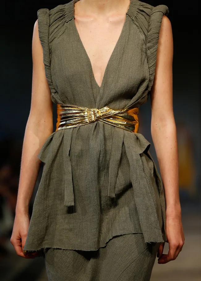 Model wearing a sleeveless olive dress with a deep v-neckline and a shiny gold belt on a fashion runway.