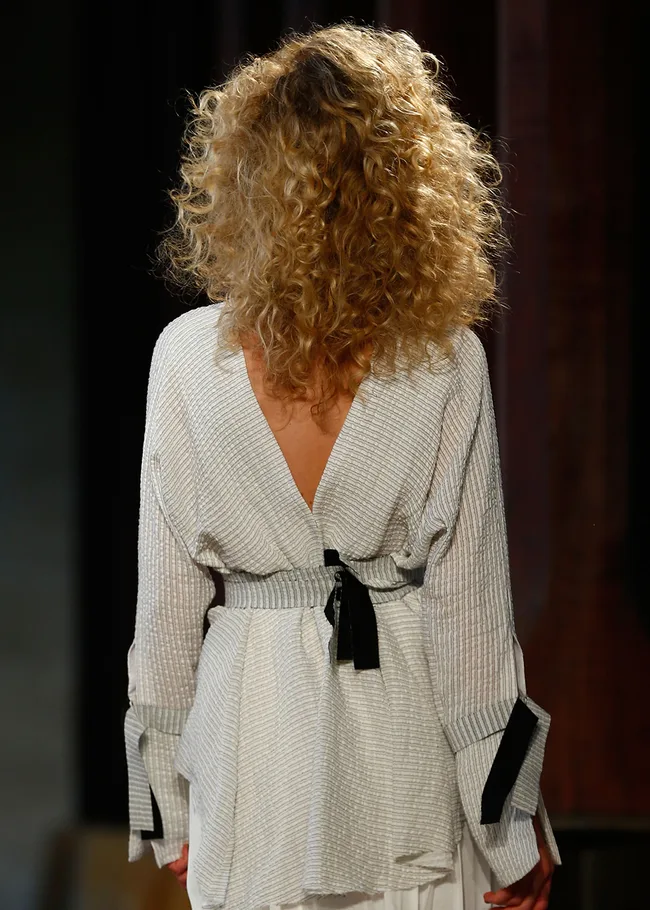 Back view of a person with curly blonde hair wearing a white textured top with black accents.