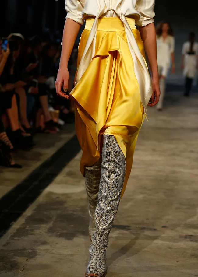 Model in yellow skirt and grey snake-print boots on runway. White top tied at waist. Fashion show setting.
