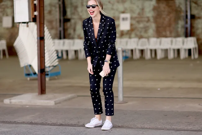 Smiling woman in a navy patterned suit and white sneakers, standing outdoors with stacked chairs in the background.