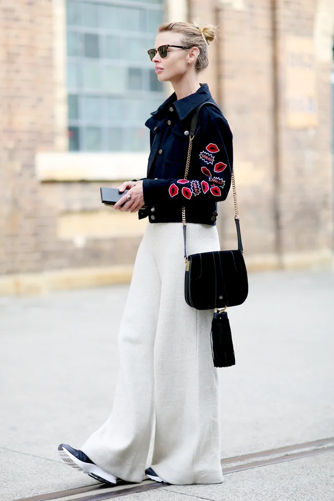 Blonde woman in sunglasses, wearing a black jacket with red designs, white pants, and a black bag, walking on a city street.