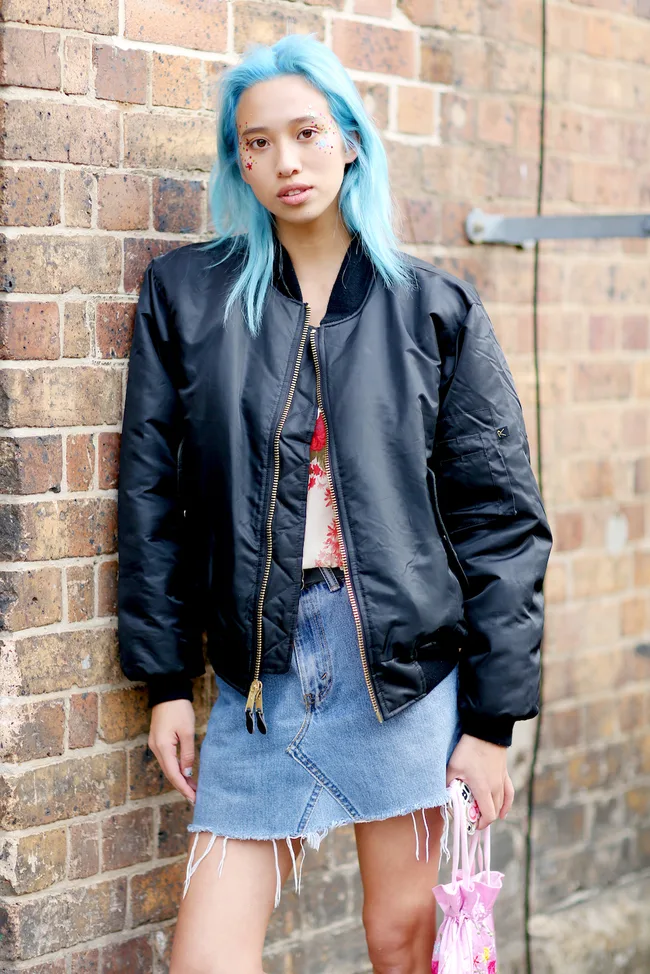 Person with blue hair and star makeup wears a black jacket and denim skirt, standing against a brick wall.