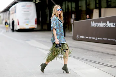 Woman in a floral shirt and green skirt with fringe, wearing heels, sunglasses, carrying a camera, walking on a street.