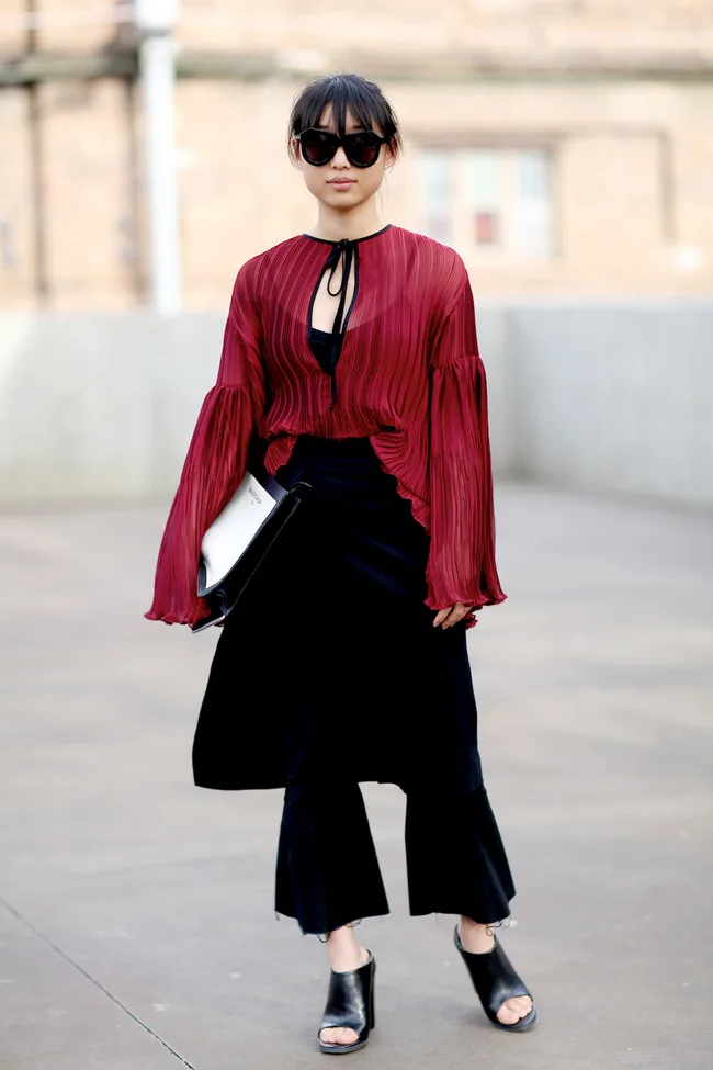 A person in sunglasses wearing a red pleated blouse, black pants, and open-toe shoes, holding a clutch.