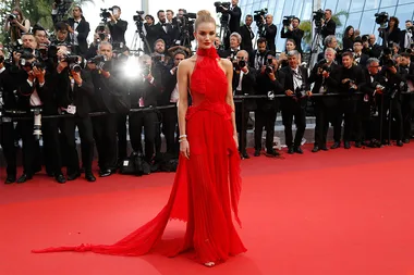 Model in a flowing red gown on a red carpet, surrounded by photographers.