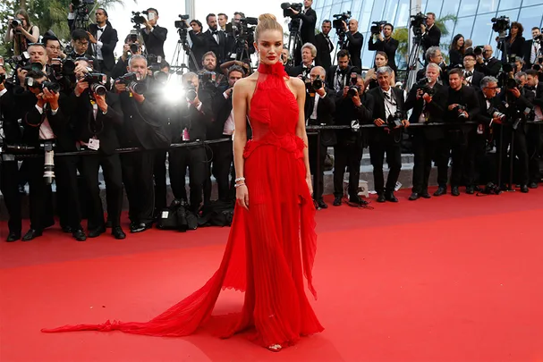 Model in a flowing red gown on a red carpet, surrounded by photographers.