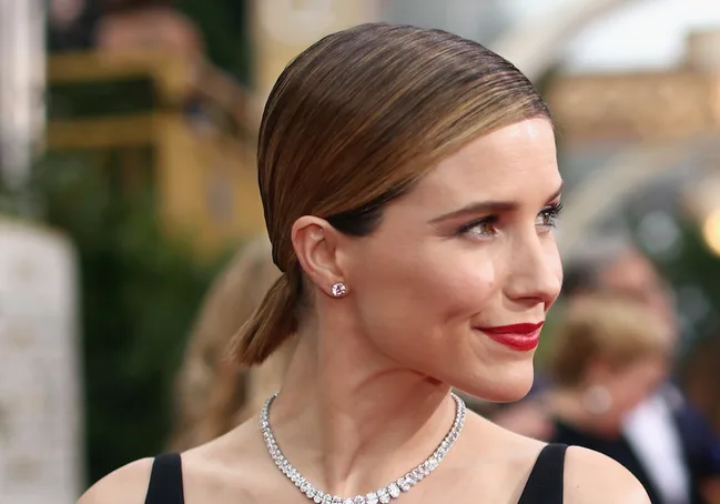 A woman with slicked-back hair, wearing a diamond necklace and red lipstick at a formal event, looking to her right.