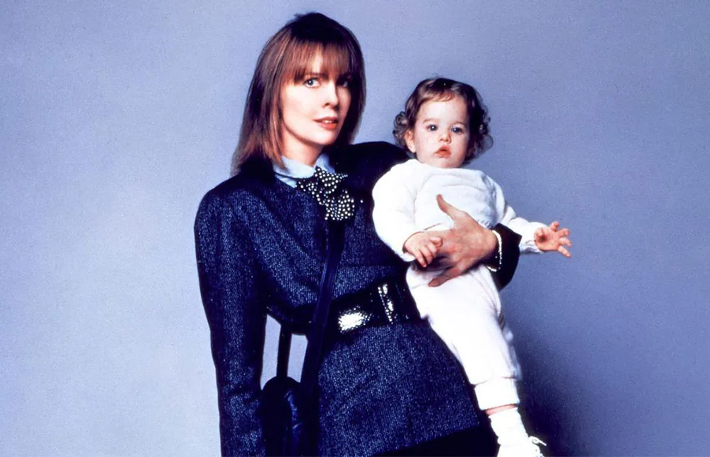 Woman in a dark suit holds a baby in white against a plain backdrop.