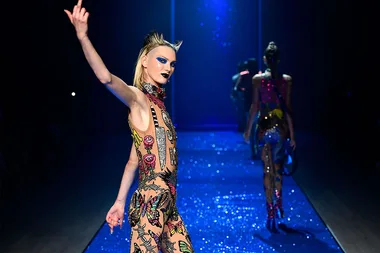 Model walking on a glittery runway, wearing a colorful, embellished bodysuit with bold makeup and hairstyle.