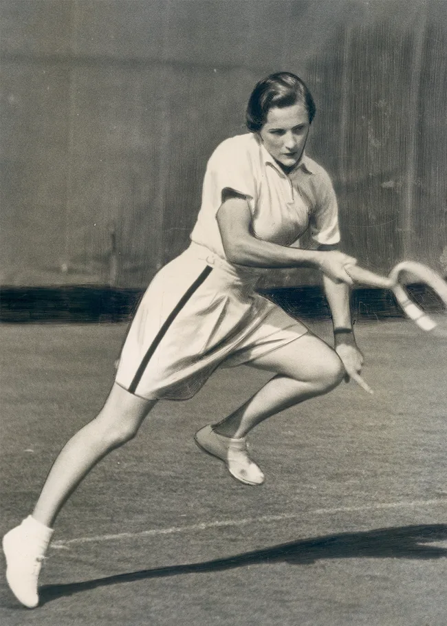 A woman playing tennis in vintage attire, caught mid-action on a grass court, holding a racquet.
