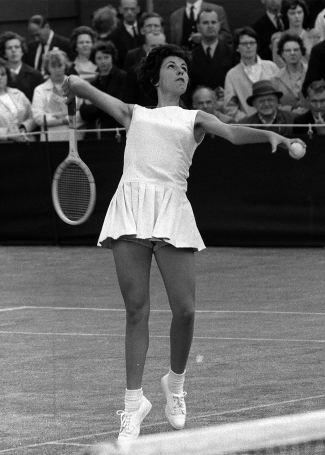 A female tennis player in mid-serve on a grass court, with an audience watching in the background.