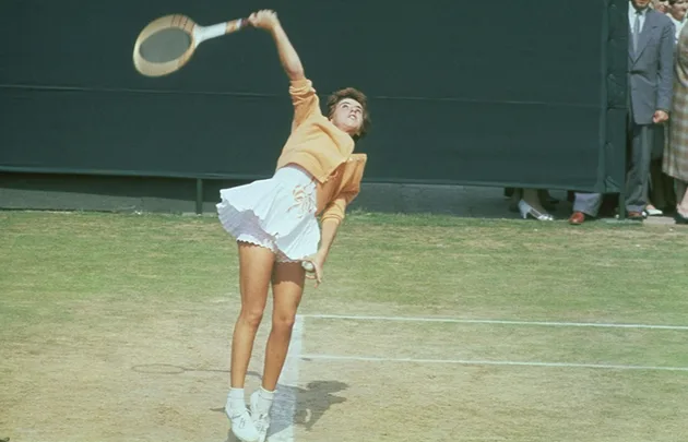 A tennis player in mid-air motion, serving on a grass court, dressed in orange and white.