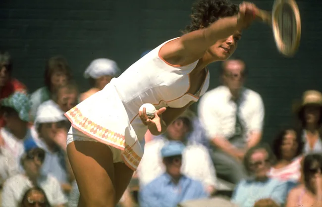 Female tennis player serving at a tournament, wearing a white outfit. Spectators watch in the background.