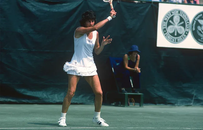 Tennis player in white dress swinging racket on outdoor court with dark backdrop and spectators in background.