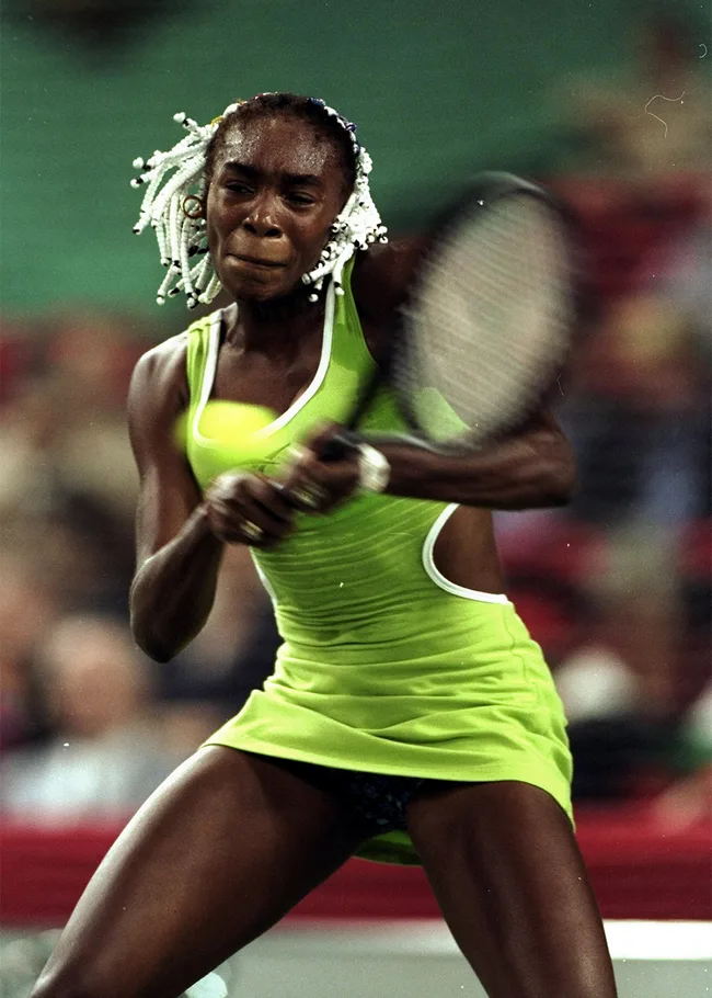 Tennis player in action, wearing a green outfit and white beaded braids, focused on returning a shot.