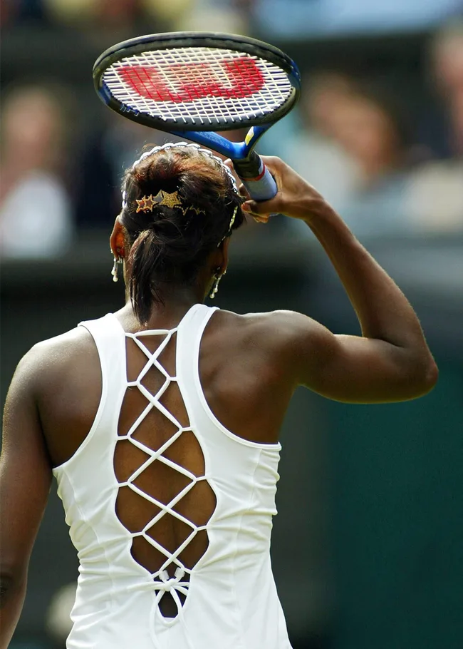 A tennis player in a white lace-up dress holds a racket, preparing to serve at Wimbledon.