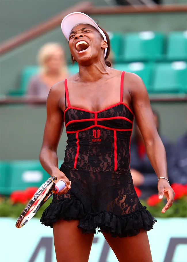 Tennis player wearing a black and red lace outfit, holding a racket, with mouth open, appearing to shout on the court.