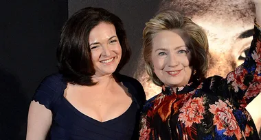 Two women posing together, one winking, wearing a navy dress, the other smiling, wearing a floral outfit.