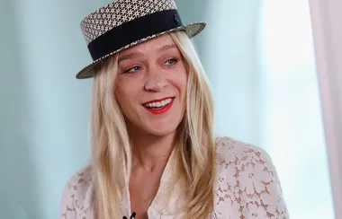 A person with long blonde hair and a patterned hat smiles, wearing a lace top.