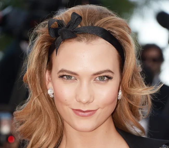 A person with blonde hair and a black headband smiles at the camera, wearing diamond earrings.