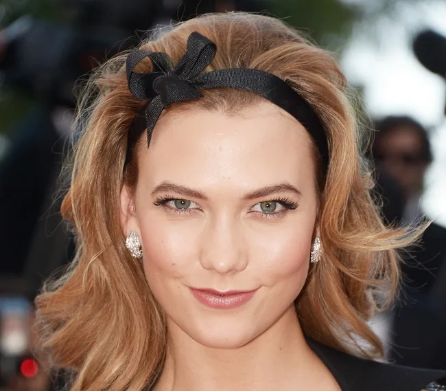 A person with blonde hair and a black headband smiles at the camera, wearing diamond earrings.