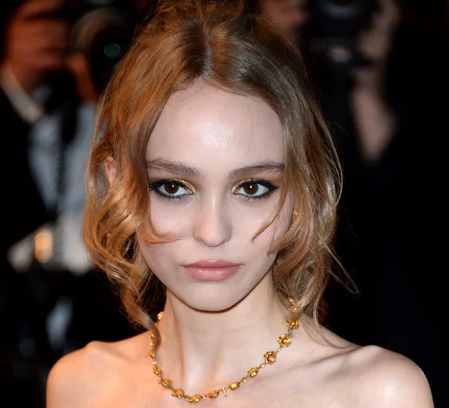 Young woman with wavy hair and a gold necklace at an event, surrounded by photographers.