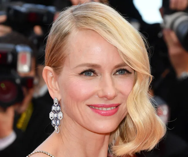 A person with blonde hair and gemstone earrings smiling, surrounded by cameras.