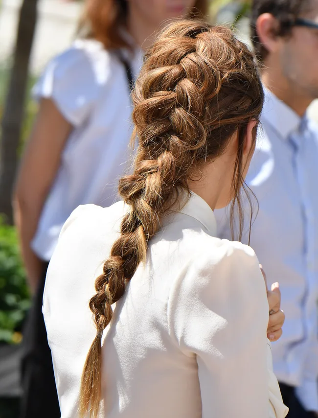 Back view of a person with a long, thick braided hairstyle, wearing a cream-colored blouse in a sunny outdoor setting.