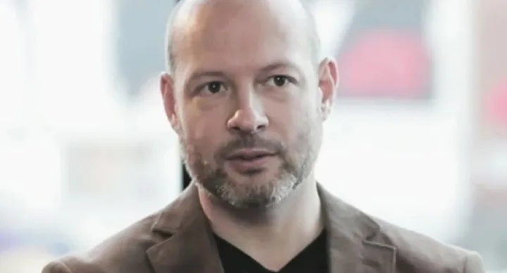 A man with a short beard and bald head wearing a brown jacket and black shirt, looking serious, in a blurred background.
