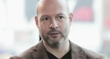 A man with a short beard and bald head wearing a brown jacket and black shirt, looking serious, in a blurred background.