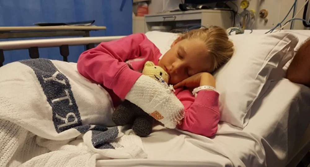 A child in a hospital bed, wearing a pink top, hugging a toy and resting peacefully.