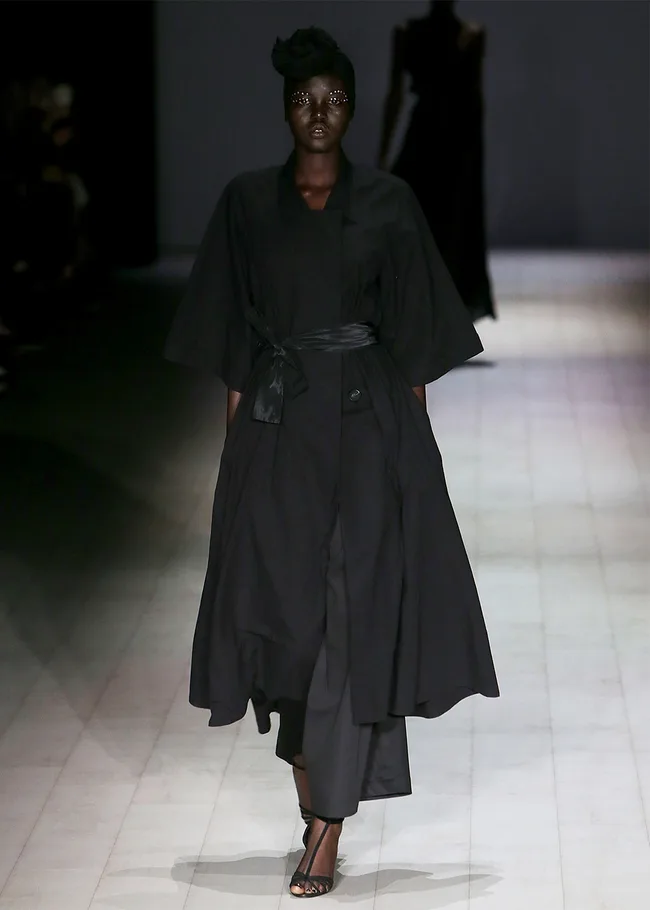 Model in a black outfit walks runway with dramatic eye makeup and hairpiece at a fashion show.