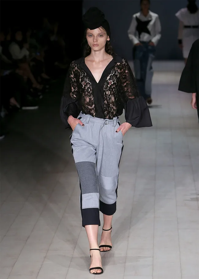 Model in a black lace top and gray patchwork pants walks the runway, wearing a headpiece and sandals.